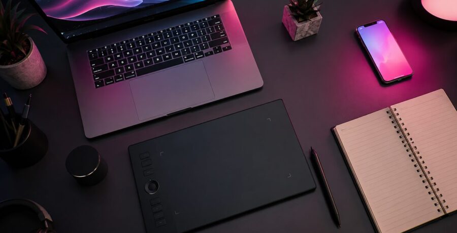 Mesa de trabalho com laptop, tablet de desenho e celular com luz rosa-magenta, representando ferramentas de criação